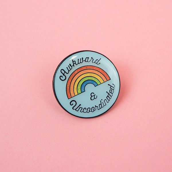 Round, light blue pin with a glitter rainbow in the centre that reads Awkward & Uncoordinated. The pin has a glossy, epoxy layer over the surface.