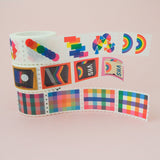 Stack of three rolls of washi tape in a stamp style with perforated edges. Each stamp has a different, colourful design. One roll has rainbow gingham stamps, one has retro VHS tapes, and the third has rainbow-coloured shapes.