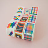 Three rolls of washi tape in a stamp style with perforated edges. Each stamp has a different, colourful design. One roll has rainbow gingham stamps, one has retro VHS tapes, and the third has rainbow-coloured shapes.