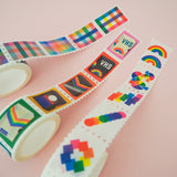 Three rolls of washi tape in a stamp style with perforated edges. Each stamp has a different, colourful design. One roll has rainbow gingham stamps, one has retro VHS tapes, and the third has rainbow-coloured shapes.