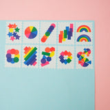 Ten stamps cut from a roll of washi tape with perforated edges. Each stamp has a different design featuring rainbows and brightly coloured shapes. They are arranged in two rows of five on a pale blue card.