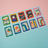 Ten stamps cut from a roll of washi tape with perforated edges. Each stamp has a different retro VHS tape design with a colourful bacground. They are arranged in two rows of five on a pale blue card.