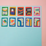 Ten stamps cut from a roll of washi tape with perforated edges. Each stamp has a different retro VHS tape design with a colourful bacground. They are arranged in two rows of five on a pale blue card.