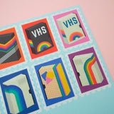 Close up of stamps cut from a roll of washi tape with perforated edges. Each stamp has a different retro VHS tape design with a colourful bacground. They are arranged in two rows on a pale blue card.