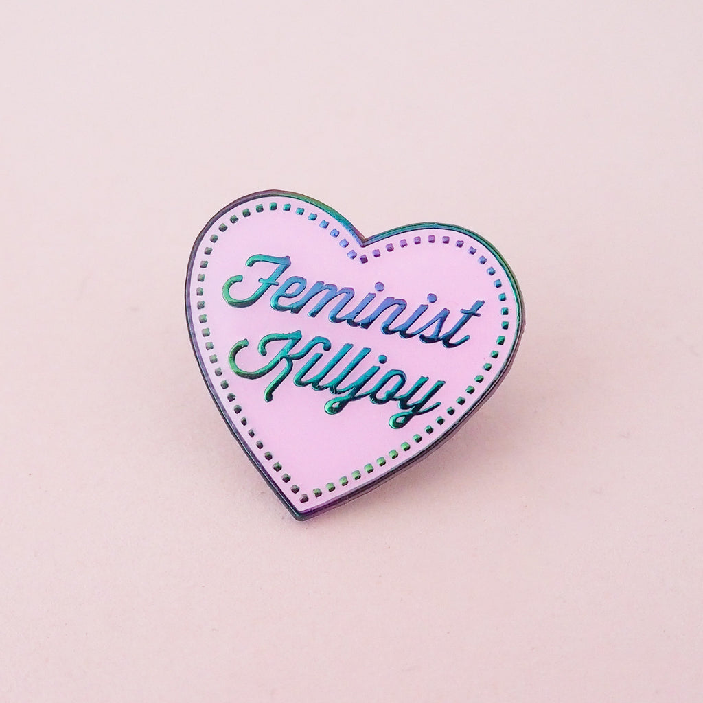 A pink enamel pin in the shape of a heart. The words Feminist Killjoy are written in rainbow-plated metal in a flowing script. Rainbow-plated dots run along the inside border of the heart. The pin is shown on a pink background.
