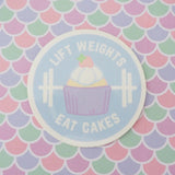 Round vinyl sticker depicting a pastel-coloured cupcake and barbell on a pale blue background. The words Lift Weights Eat Cakes are written in white capitals. The sticker has a white border.