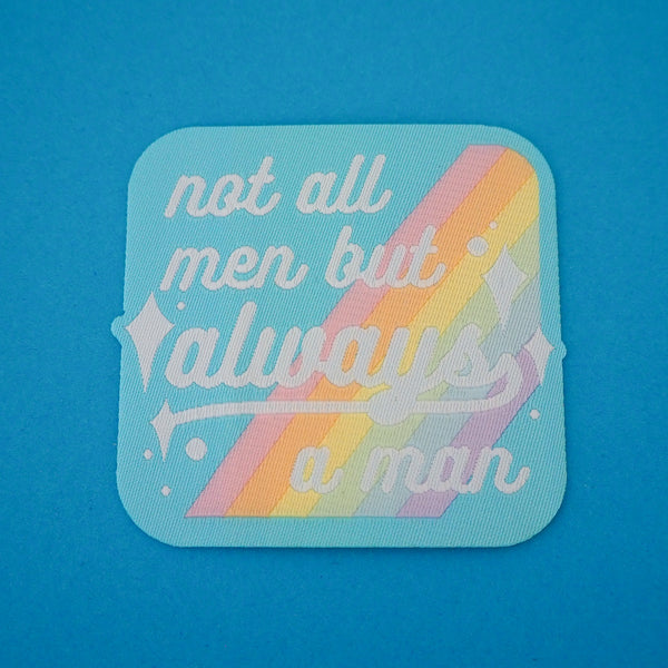 Square woven patch with rounded corners that reads not all men but always a man in white script with decorative white sparkles. The background of the patch is pale blue with pastel rainbow stripes running diagonally from top-right to bottom-left. The patch has a pale blue border.