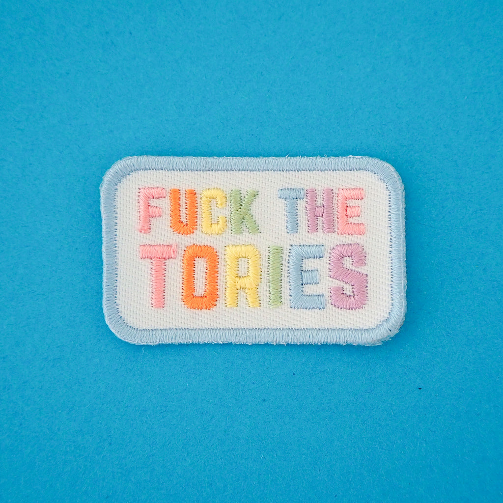 Rectangular, white embroidered patch with rounded corners. It reads Fuck the Tories in a rainbow of pastel coloured capital letters. The patch has pastel blue merrowed edges.