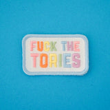 Rectangular, white embroidered patch with rounded corners. It reads Fuck the Tories in a rainbow of pastel coloured capital letters. The patch has pastel blue merrowed edges.