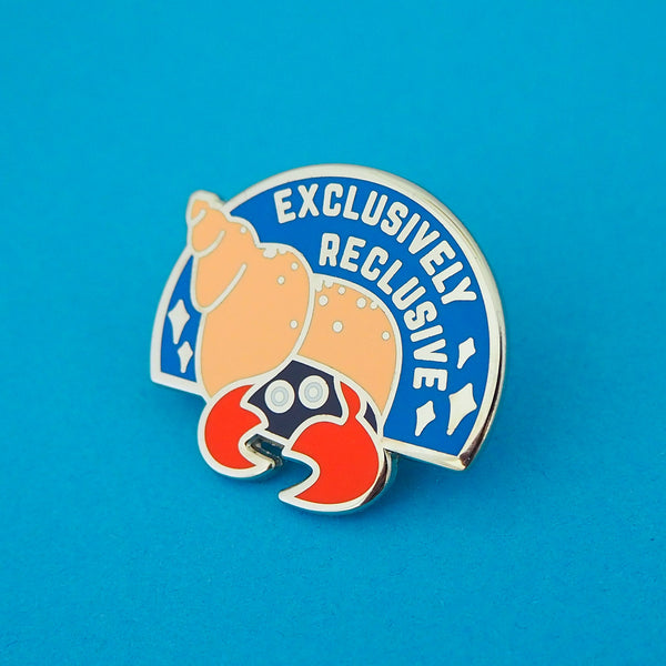 Semi-circular, blue enamel pin with the image of a hermit crab in a peach coloured spiral shell. Only the crabs eyes and red claws are visible, peeking out from the darkness of the shell's entrance. The words Exclusively Reclusive are written on the blue section in silver capital letters with some silver sparkles. The pin is on a blue background.