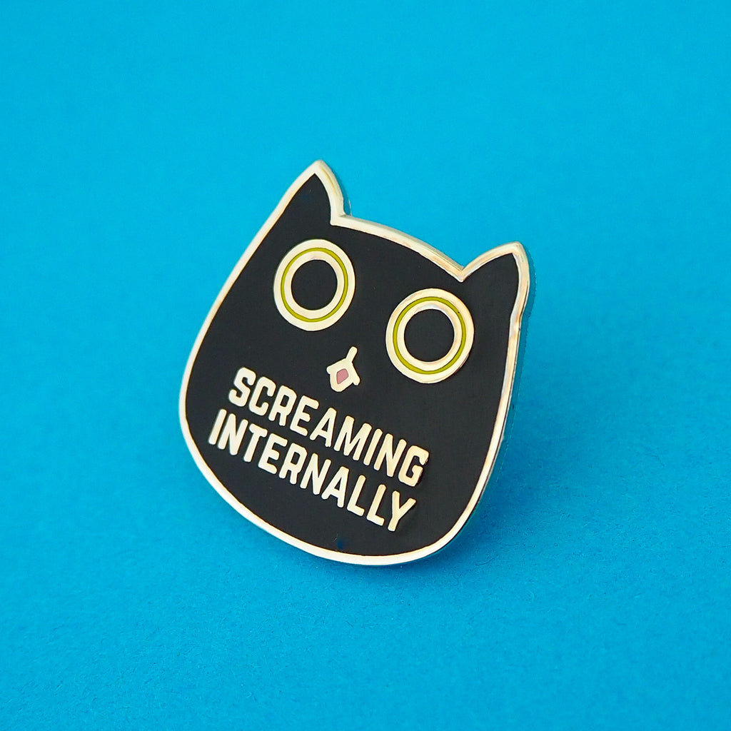 Metal pin showing a black cat head with bright green eyes. The pupils are large and round and its tongue is poking out. Beneath its mouth are the words Screaming Internally in silver capital letters.