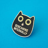Metal pin showing a black cat head with bright green eyes. The pupils are large and round and its tongue is poking out. Beneath its mouth are the words Screaming Internally in silver capital letters.