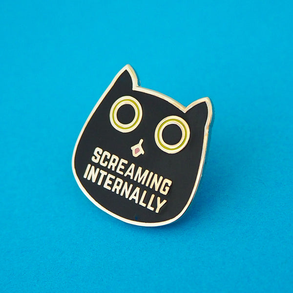 Metal pin showing a black cat head with bright green eyes. The pupils are large and round and its tongue is poking out. Beneath its mouth are the words Screaming Internally in silver capital letters.