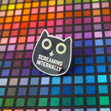 Metal pin showing a black cat head with bright green eyes. The pupils are large and round and its tongue is poking out. Beneath its mouth are the words Screaming Internally in silver capital letters.