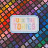 Rectangular, white embroidered patch with rounded corners. It reads Fuck the Tories in a rainbow of pastel coloured capital letters. The patch has pastel blue merrowed edges.