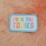 Rectangular, white embroidered patch with rounded corners. It reads Fuck the Tories in a rainbow of pastel coloured capital letters. The patch has pastel blue merrowed edges.