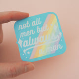 Square woven patch with rounded corners that reads not all men but always a man in white script with decorative white sparkles. The background of the patch is pale blue with pastel rainbow stripes running diagonally from top-right to bottom-left. The patch has a pale blue border and is held between a finger and thumb.
