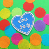Heart-shaped, light-blue patch. In the centre of the heart are the words Boss Lady embroidered in silver script. The heart is outlined with a row of tiny silver squares. The patch has silver merrowed edges.