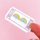 A pair of enamel stud earrings depicting pastel coloured rainbows. The earrings are on a white carnival ticket which is held between two fingers.