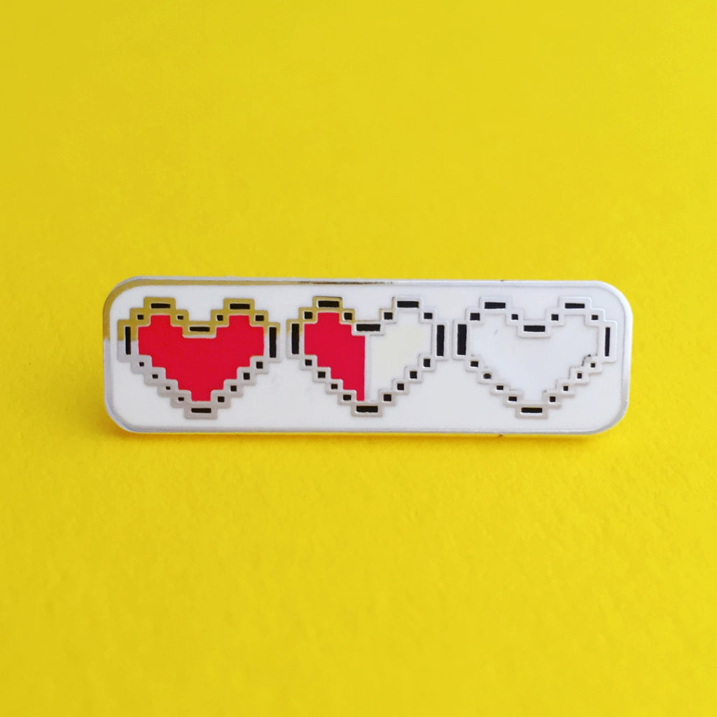 Rectangular enamel pin showing three pixelated hearts. The first heart is filled red, the second is filled red on the left half and white on the right, the third heart is filled white. The pin is on a yellow background.