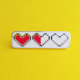 Rectangular enamel pin showing three pixelated hearts. The first heart is filled red, the second is filled red on the left half and white on the right, the third heart is filled white. The pin is on a yellow background.
