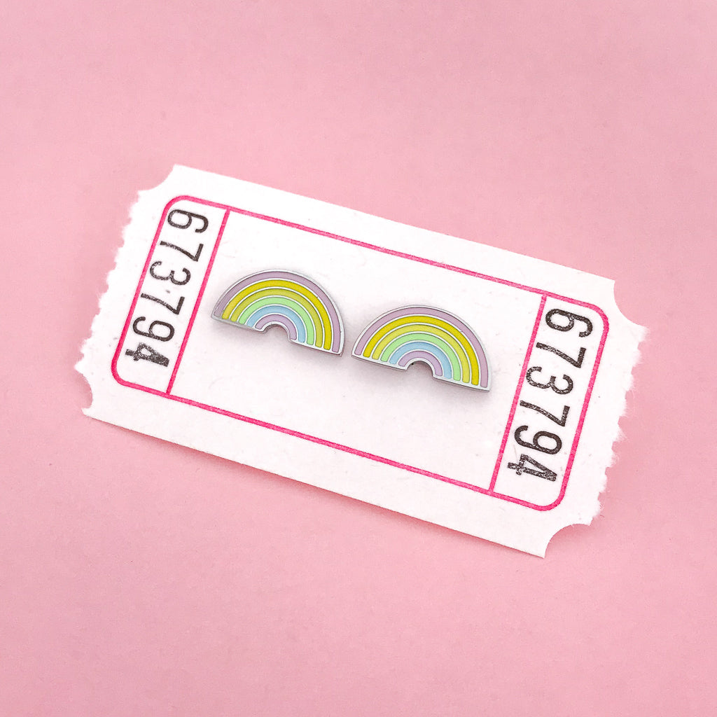 A pair of enamel stud earrings depicting pastel coloured rainbows. The earrings are on a white carnival ticket.