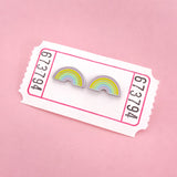 A pair of enamel stud earrings depicting pastel coloured rainbows. The earrings are on a white carnival ticket.