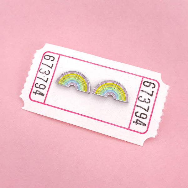 A pair of enamel stud earrings depicting pastel coloured rainbows. The earrings are on a white carnival ticket.