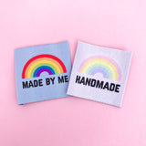 Two clothing labels. The first is pale blue with the words made by me in black capital letters and a bright rainbow above. The second is white with the word handmade in black capital letters with a pastel rainbow above. They are on a pastel pink background.