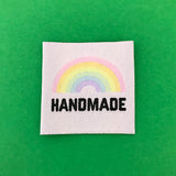 White clothing label with the word handmade in black capital letters with a pastel rainbow above. Label is on a green background.