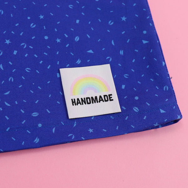 White clothing label with the word handmade in black capital letters with a pastel rainbow above.  Label is on patterned blue material.