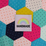 White clothing label with the word handmade in black capital letters with a pastel rainbow above. Th label is on a patchwork background.