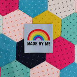 Pale blue clothing label with the words made by me in black capital letters and a bright rainbow above. The label is on a patchwork background.
