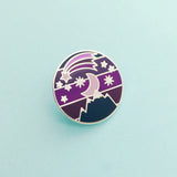 Round metal pin showing a moon and stars over a mountain on a striped background. Above the moon there is a shooting star with a long tail. The colours are a variety of dark blues and purples.
