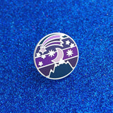 Round metal pin showing a moon and stars over a mountain on a striped background. Above the moon there is a shooting star with a long tail. The colours are a variety of dark blues and purples. The pin is on a blue glitter background.