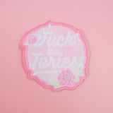 Circular, pastel pink woven patch with the words Fuck the Tories in a white script. The patch is decorated with pink roses, green foliage and white sparkles, all bursting over the edge of the circle. The patch has pink merrowed edges.