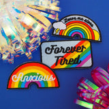 Three brightly coloured patches reading Forever Tired, Leave Me Alone, and Anxious. They are shown with rainbow rosette decorations..