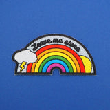 Embroidered patch showing a rainbow with sun and lightning cloud in a pale blue sky that reads Leave Me Alone in black script. The patch has a black merrowed edge.