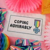 Cross stitch surrounded by decorations. Cross stitch design features a pastel coloured medal and the words Coping Admirably in bold, black letters.