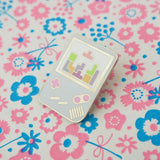 Enamel pin shaped like a light blue Gameboy with pink buttons. The white screen shows a game of tetris in progress with pastel coloured tetrominoes.