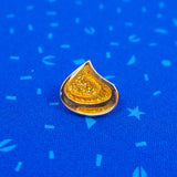 A gold glitter enamel pin with gold-coloured plating in the shape of a korok seed. The pin is on a blue patterned material.
