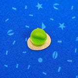 The back of a gold-coloured enamel pin with a green rubber backing. The pin is on a blue patterned material.