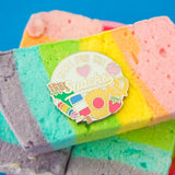 A round enamel pin with a white background showing a selection of sweets and snacks which are bursting over the edge. The colours of the snacks are muted. At the top of the pin are the words 'Here for the' in capital letters and then the word 'Snacks' in large, underlined script below a pastel pink heart. The pin is sitting on top of a pile of rainbow marshmallow.