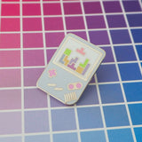 Enamel pin shaped like a light blue Gameboy with pink buttons. The white screen shows a game of tetris in progress with pastel coloured tetrominoes.