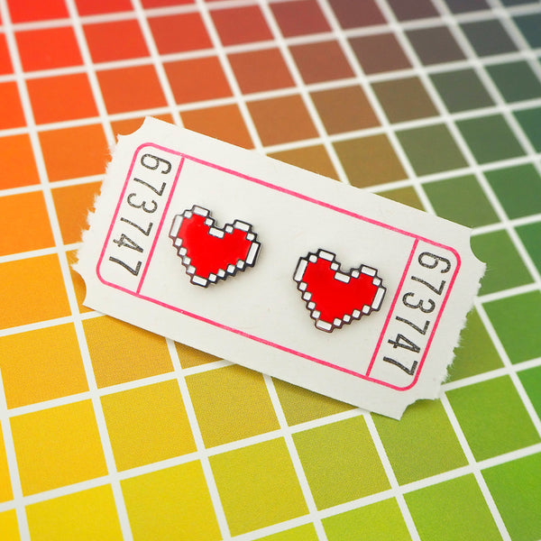 A pair of enamel stud earrings depicting a red, pixelated heart with a white outline. The earrings are on a white carnival ticket.