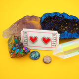 A pair of enamel stud earrings depicting a red, pixelated heart with a white outline. The earrings are on a white carnival ticket, nestled amongst a cluster of crystals.