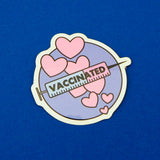 Round vinyl sticker showing a syringe with a stream of pink hearts behind it. The background is purple and the syringe is pink and blue with the word Vaccinated in black capitals. The sticker has a white border.