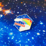 Hexagonal metal pin depicting a moon and dark blue starry sky. A white rocket flies past with a rainbow trail. A red banner near the bottom reads Intergalactic. The pin is on a starry background.