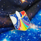 Metal pin in a shield shape. A white rocket ascends through the middle of the pin between a red ringed-planet and a white moon. The rocket leaves a rainbow trail that fans out to the bottom with the words Rocket Science flanking it.