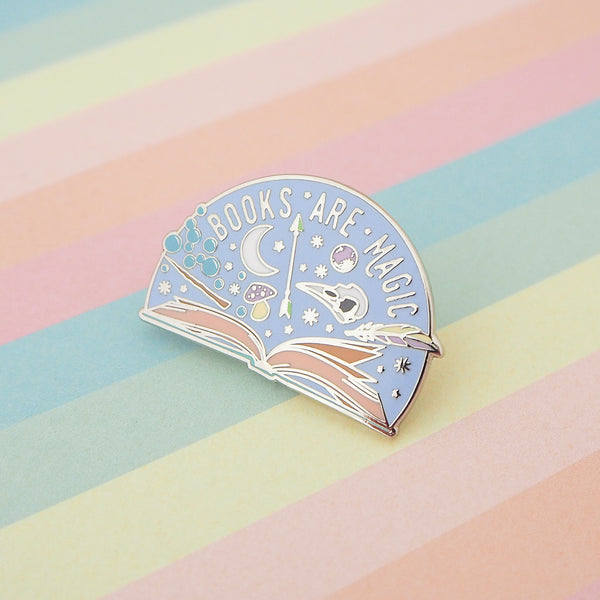 Semi-circular, lilac pin with the words Books Are Magic in capital letters. There is an open book at the bottom and many magical items are bursting forth from the pages, including a wand, raven skull, quill, stars and bubbles.  Pin is on a pastel striped background.
